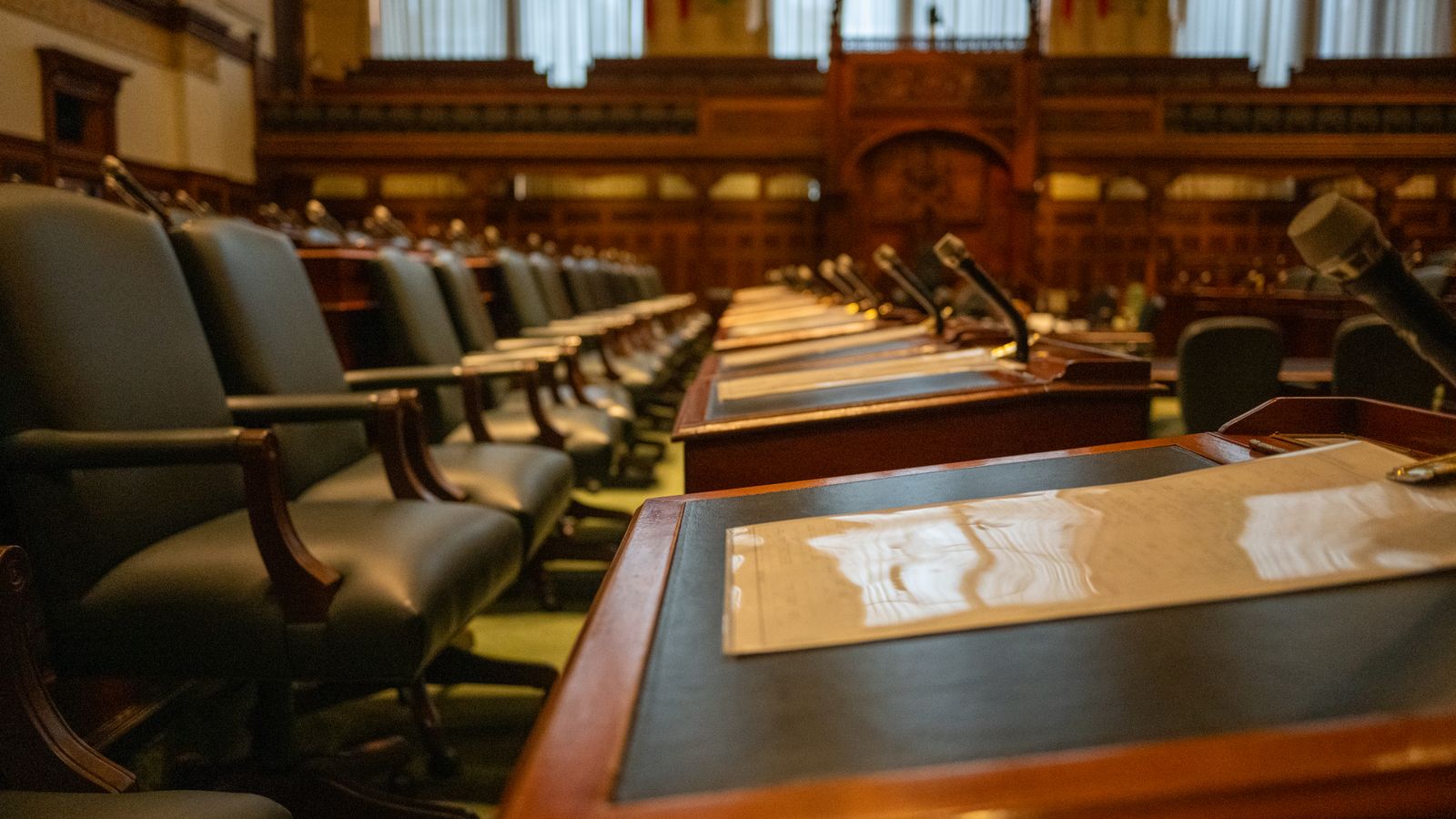 Chambre institutionnelle vide avec rangées de bureaux et microphones, évoquant la gouvernance publique